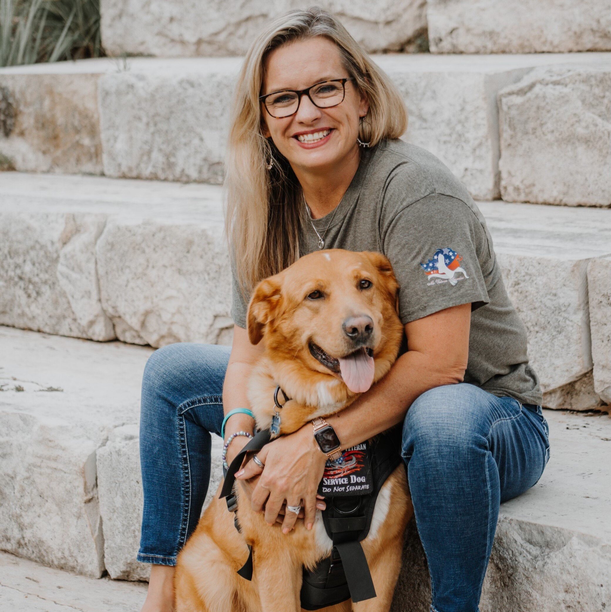 Smiling woman sitting on stone steps, cuddling a golden retriever service dog wearing a vest, showcasing a warm and friendly bond.